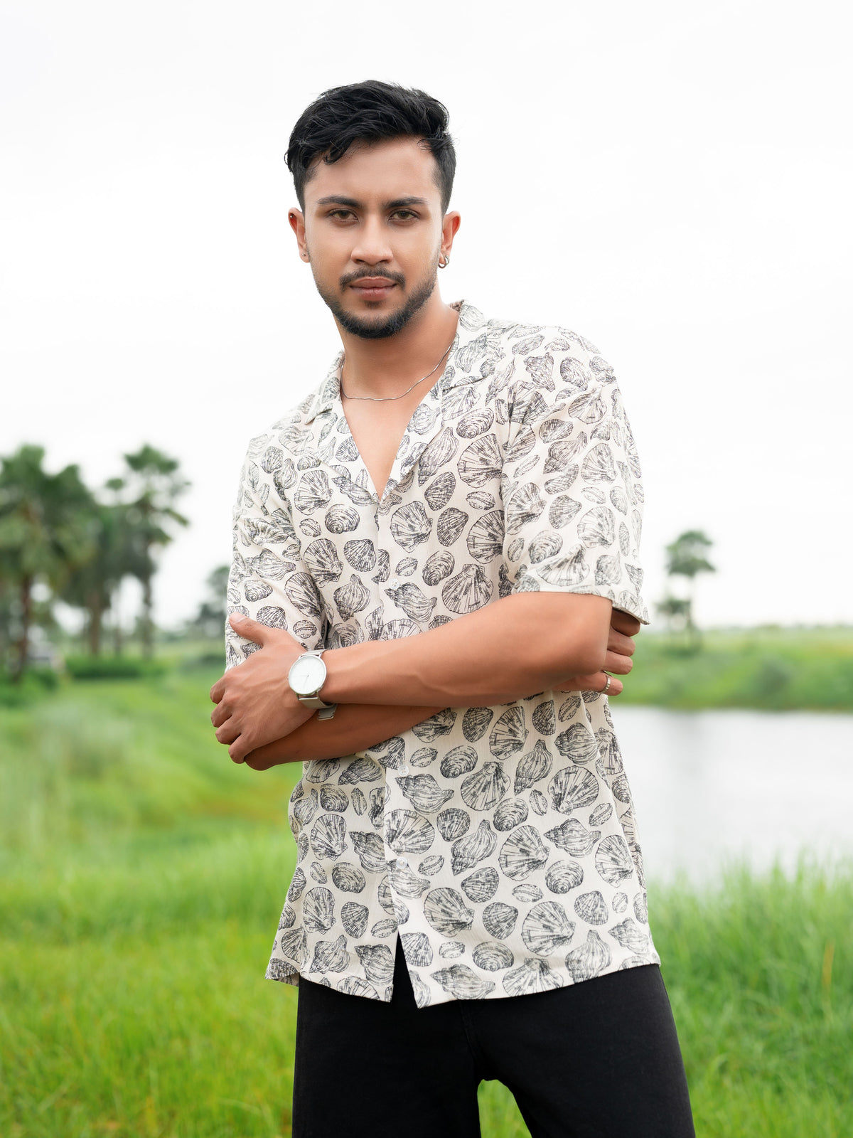 A man wearing a short-sleeve button-up shirt with a black and white leaf pattern, standing outdoors in a grassy field with a lake and palm trees in the background.