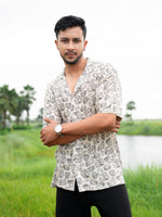 A man wearing a short-sleeve button-up shirt with a black and white leaf pattern, standing outdoors in a grassy field with a lake and palm trees in the background.